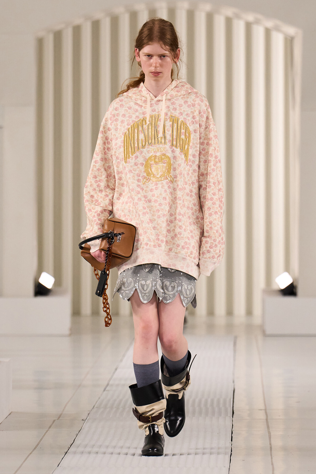 Model walking a runway in an oversized pink floral hoodie with gold lettering, a gray scalloped-hem lace skirt, gray socks, and black boots, carrying a tan purse with a chain strap.