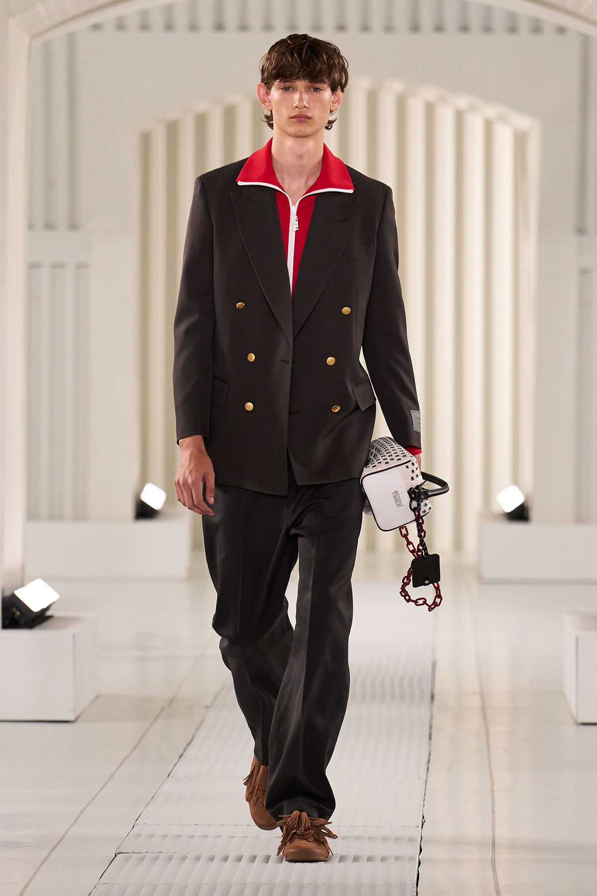 Male model walks the runway in a black double-breasted blazer with a red collar, dark trousers, and brown fringe shoes, carrying a white handbag.