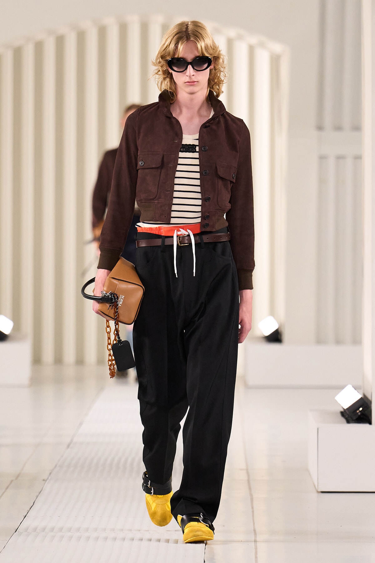Model walking runway in a brown cropped jacket over striped top, black wide-leg pants with a red belt, yellow shoes, and oversized sunglasses, carrying a tan bag.