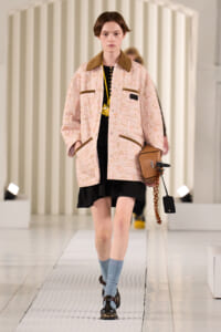 Model walking a runway in a pastel pink floral quilted jacket with brown collar and trim, over a black dress, blue socks, and brown shoes, carrying a tan handbag.