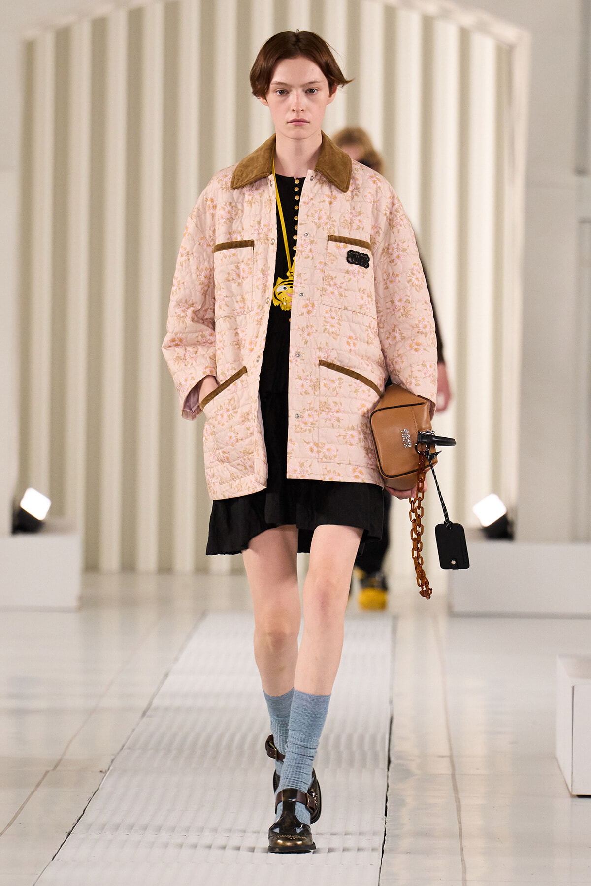 Model walking a runway in a pastel pink floral quilted jacket with brown collar and trim, over a black dress, blue socks, and brown shoes, carrying a tan handbag.