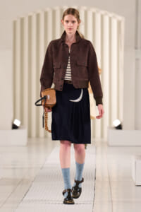Model on a runway wearing a brown cropped suede jacket, striped top, navy skirt with a crescent moon, blue socks, brown sandals, and a tan bag.