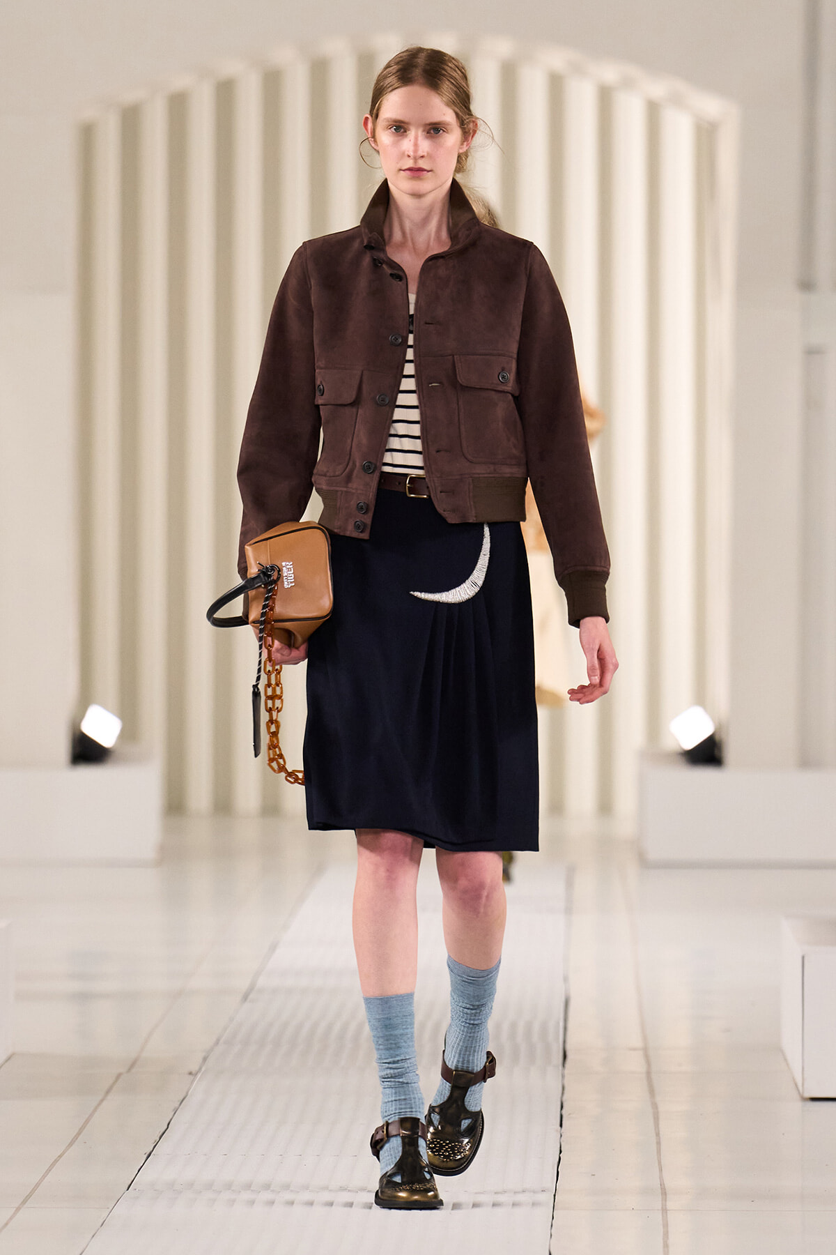 Model on a runway wearing a brown cropped suede jacket, striped top, navy skirt with a crescent moon, blue socks, brown sandals, and a tan bag.