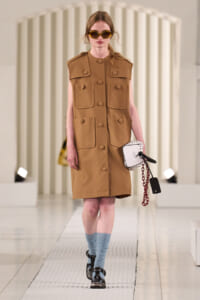 Model wearing a camel sleeveless utility coat with large pockets and tan buttons, blue socks, black shoes, and a white handbag with a chain on a runway.