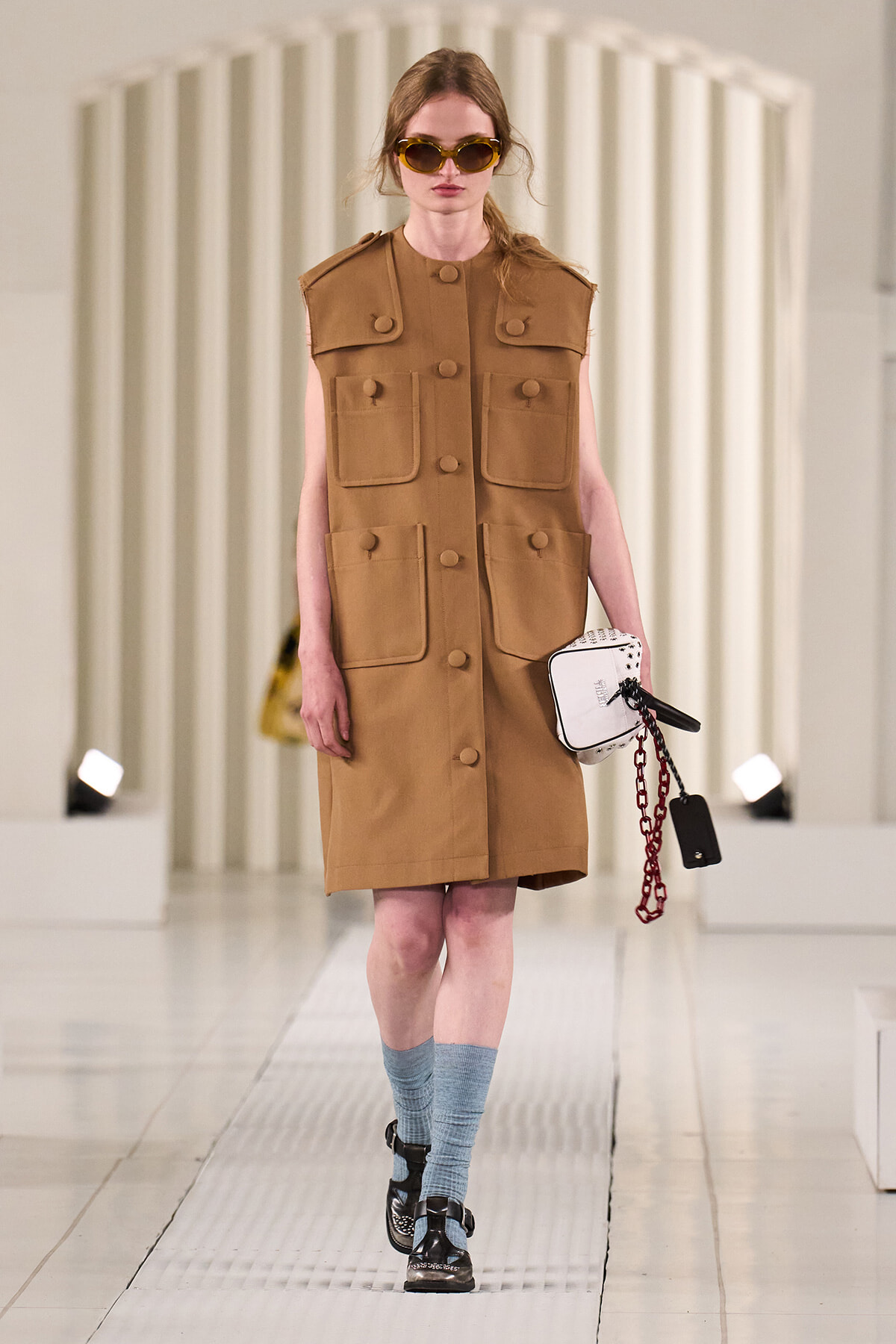 Model wearing a camel sleeveless utility coat with large pockets and tan buttons, blue socks, black shoes, and a white handbag with a chain on a runway.