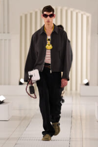 Male model on a runway wearing a black zip jacket over a striped tee, with a bold yellow lion pendant and burgundy sunglasses.