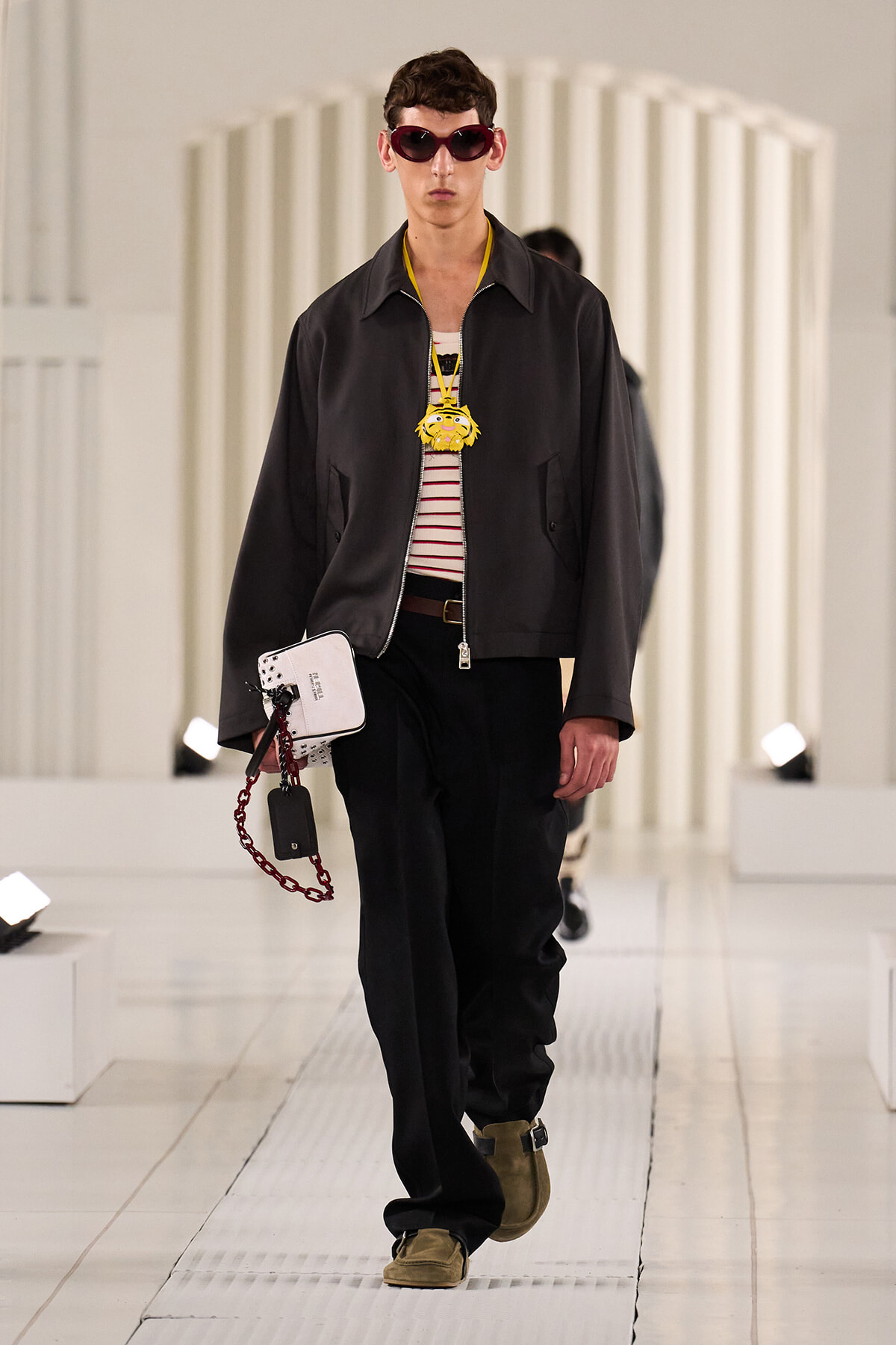 Male model on a runway wearing a black zip jacket over a striped tee, with a bold yellow lion pendant and burgundy sunglasses.