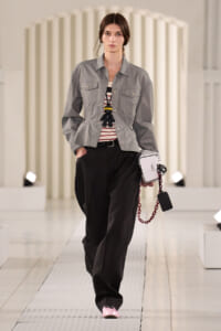 Model on a white runway wearing a gray button-front jacket over a striped top, with wide black trousers and pink shoes, carrying a white handbag with a red chain and a black tag.