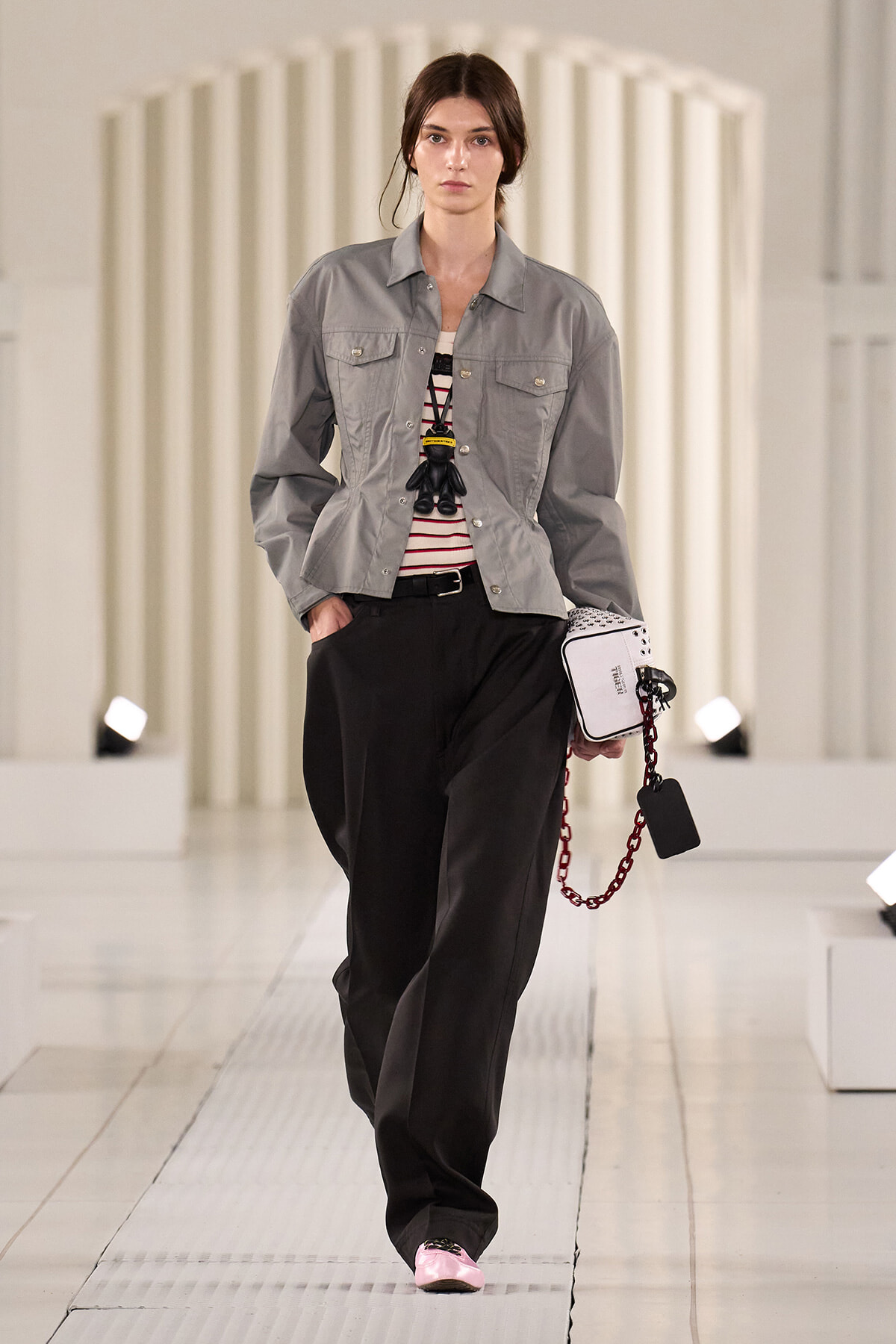Model on a white runway wearing a gray button-front jacket over a striped top, with wide black trousers and pink shoes, carrying a white handbag with a red chain and a black tag.