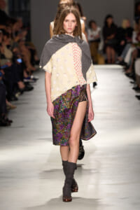 Model walking the runway in a layered outfit: cream polka-dotted top, gray shawl, and a colorful textured wrap skirt with a high slit, paired with gray leg warmers and brown shoes.