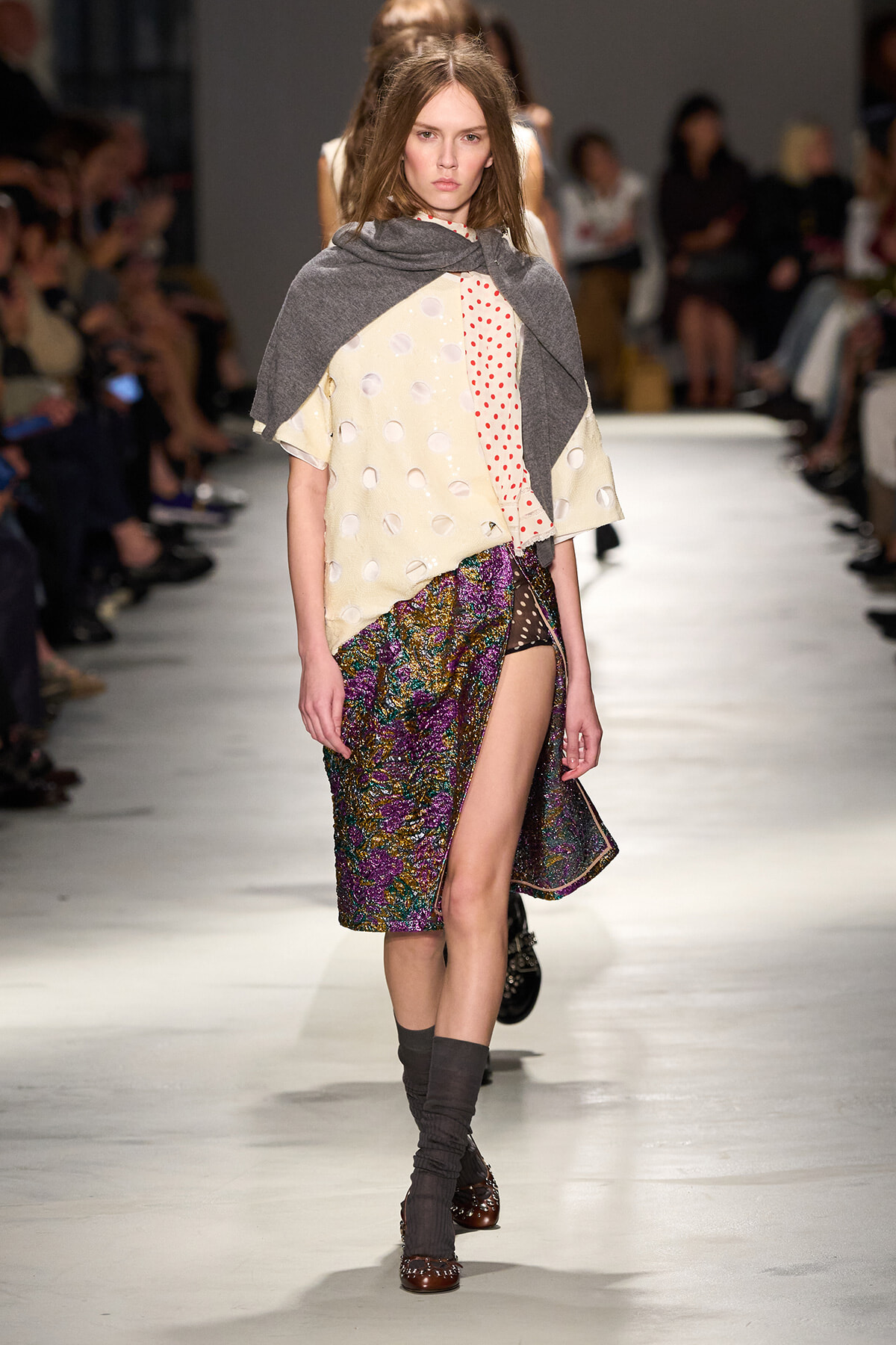 Model walking the runway in a layered outfit: cream polka-dotted top, gray shawl, and a colorful textured wrap skirt with a high slit, paired with gray leg warmers and brown shoes.