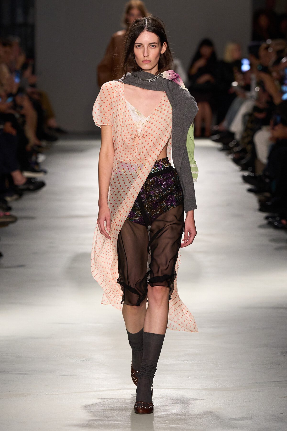 Model walks runway in a peach sheer polka-dot wrap dress over black sheer pants and iridescent shorts, paired with a gray scarf draped over one shoulder and brown shoes. Audience in background.