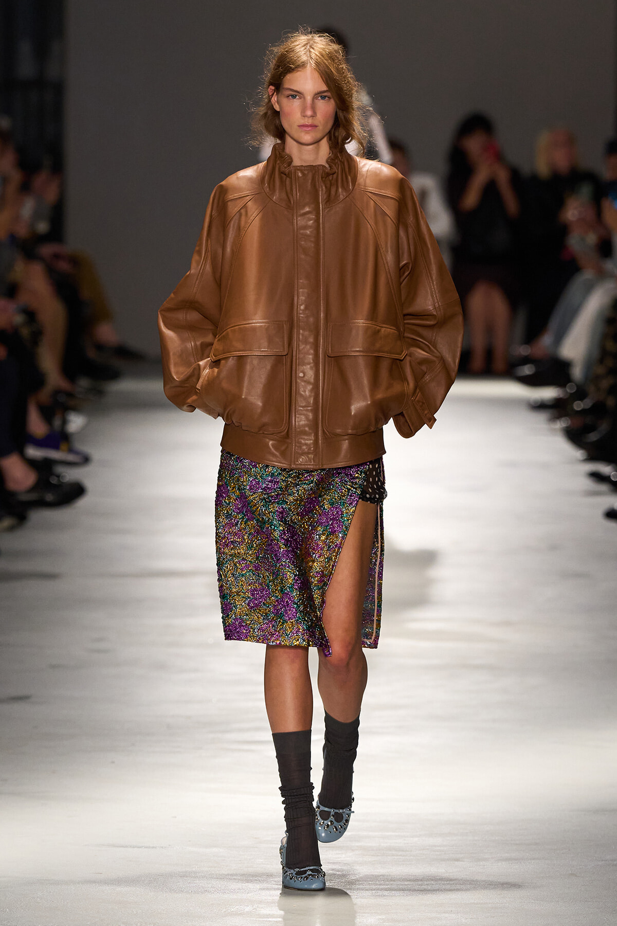 Model walking a fashion show wearing an oversized tan leather jacket and a colorful sequined skirt with a front slit, blue shoes visible at the toes, audience in the background.