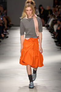 Model walking a fashion runway in a gray knit top, bright orange ruffled skirt, and a pink scarf draped over the shoulder with blue studded shoes.