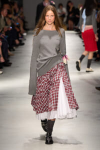 Model walking a fashion show wearing a gray knit sweater over a layered red plaid skirt with a white underskirt and a pink flower accessory at the waist. Her boots and socks complete the look.