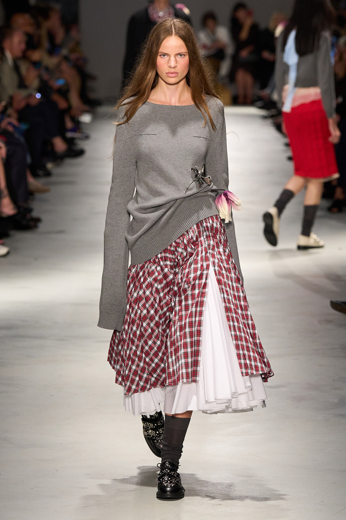 Model walking a fashion show wearing a gray knit sweater over a layered red plaid skirt with a white underskirt and a pink flower accessory at the waist. Her boots and socks complete the look.