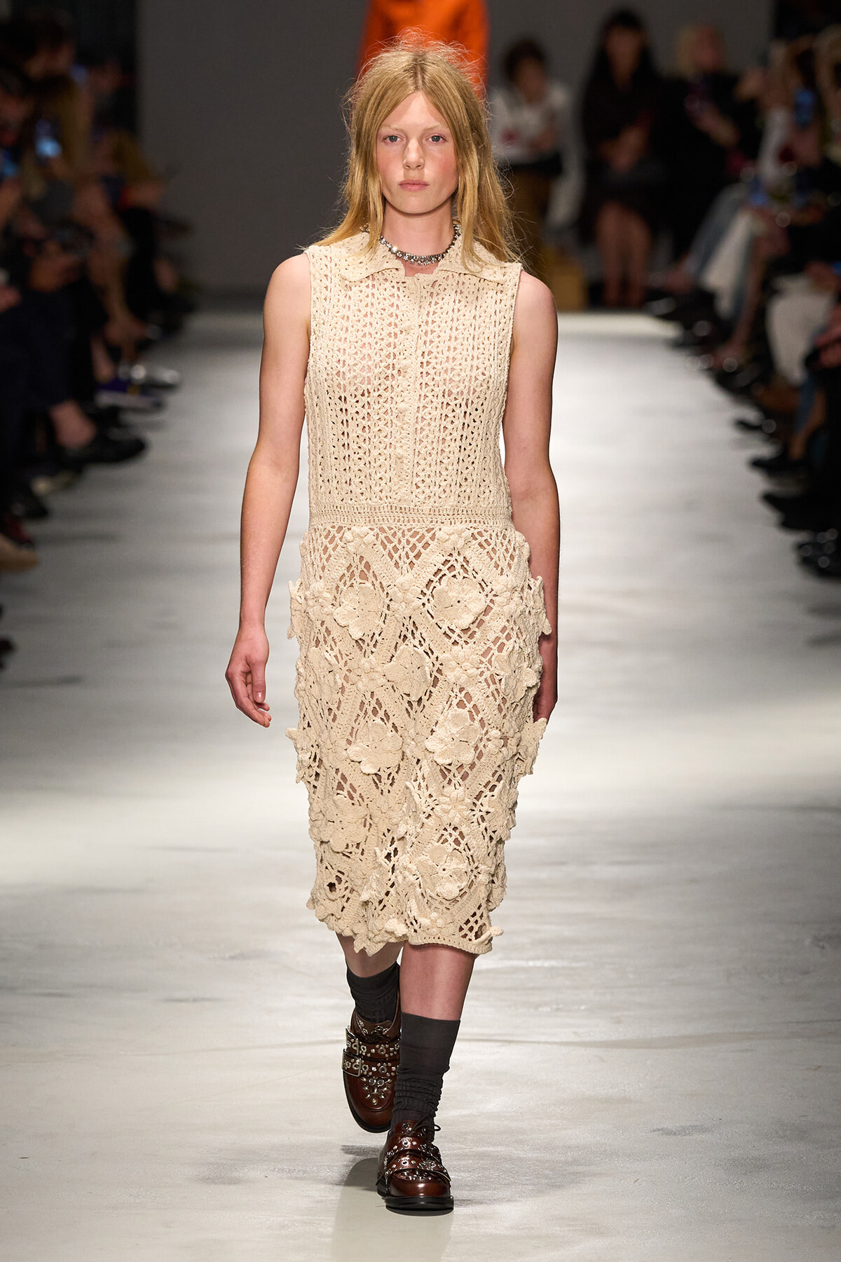 Model walking a runway in a sleeveless beige crochet lace midi dress with a collar, paired with brown loafers and dark socks. Audience out of focus in the background.3a5️