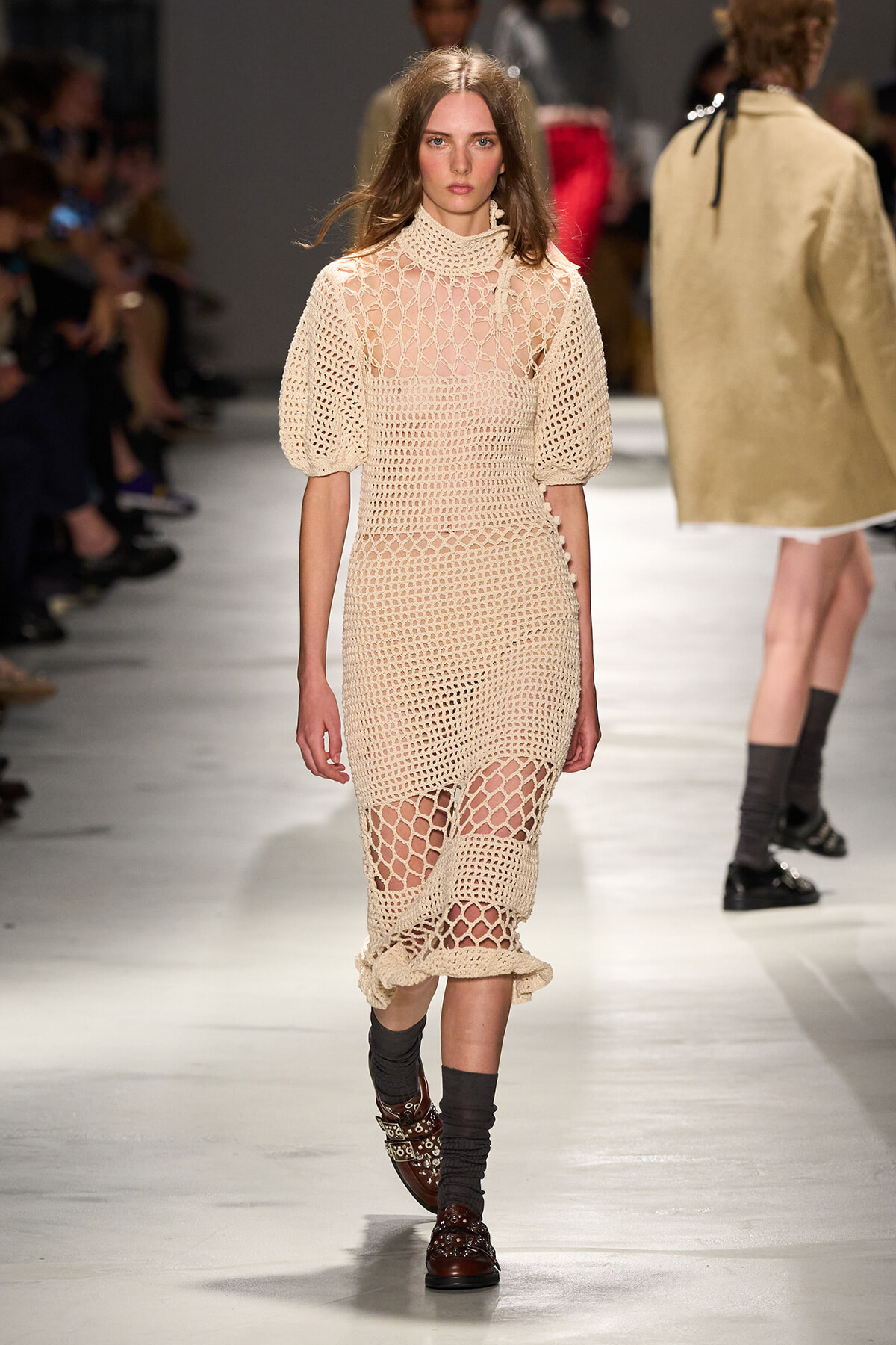 Model walking the runway in a beige crochet knit midi dress with a high neck and short puff sleeves, showing leg through openwork panels. She wears dark socks and brown studded boots.