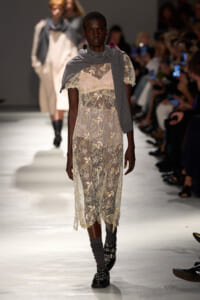 Model walking a fashion show in a sheer cream lace dress with floral embroidery and a gray scarf draped over the shoulders.
