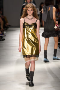 Model walks a fashion runway in a gold metallic slip dress with black lace trim at the neckline and hem, wearing a layered necklace and black boots with studs.