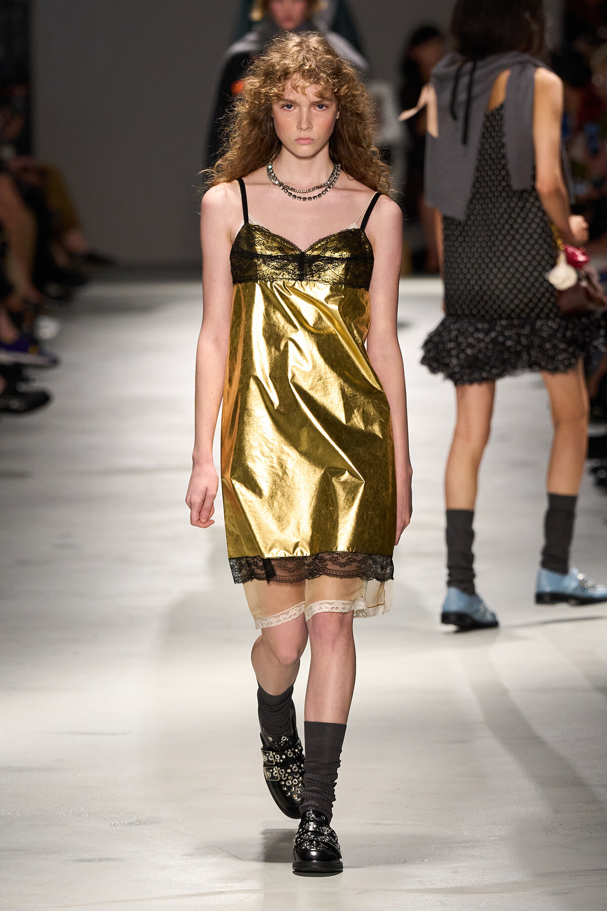 Model walks a fashion runway in a gold metallic slip dress with black lace trim at the neckline and hem, wearing a layered necklace and black boots with studs.