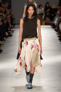 Model walking a fashion runway in a sleeveless dark brown button-up top over a floral cream skirt with lace panel, studded belt, and light blue boots on a white catwalk with spectators in the background.