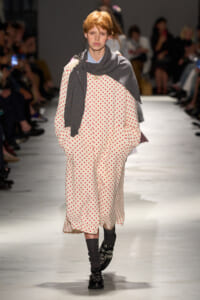 Model walking runway in a cream dress with red polka dots, draped in a gray scarf and dark accessories.