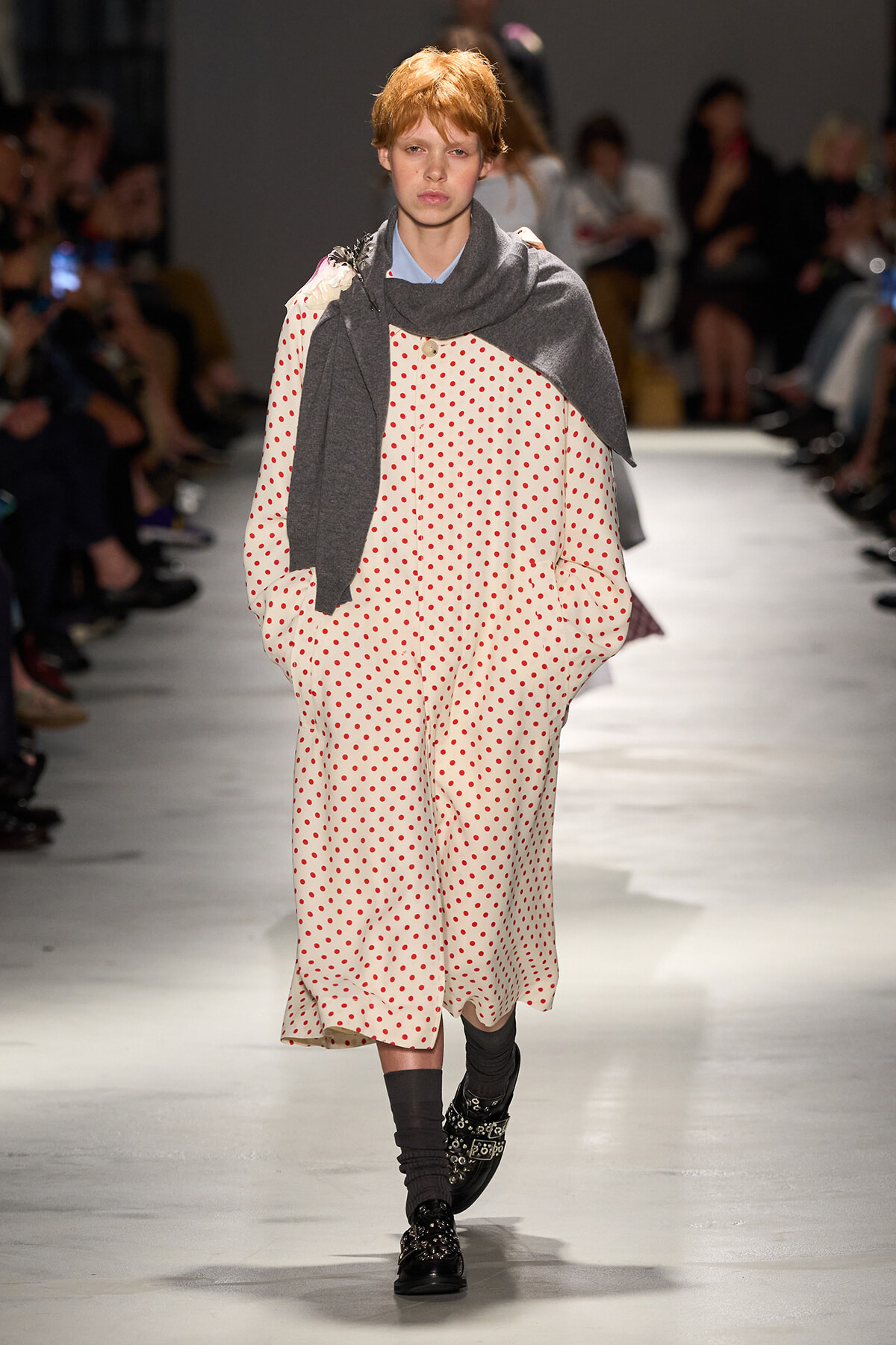 Model walking runway in a cream dress with red polka dots, draped in a gray scarf and dark accessories.