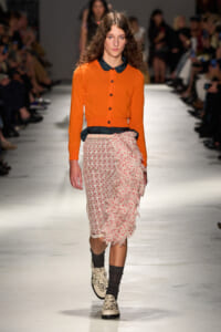 Model walks the runway in an orange cardigan with a dark collar, paired with a sheer pink polka-dot skirt and dramatic ruffle detail at the side; black socks and patterned cream boots.