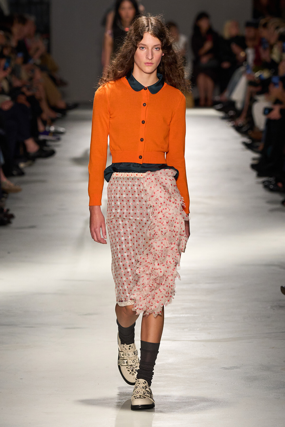 Model walks the runway in an orange cardigan with a dark collar, paired with a sheer pink polka-dot skirt and dramatic ruffle detail at the side; black socks and patterned cream boots.