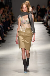 Model walking down a runway in a beige pencil skirt, lace top, and gray scarf draped over shoulders; audience blurred in background.
