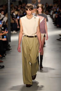 Male model in a sleeveless cream textured top and wide khaki trousers walks the runway at a fashion show with an audience in the background.