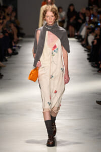 Female model walks the runway in a cream satin dress with floral embroidery, a gray cape, and an orange clutch.