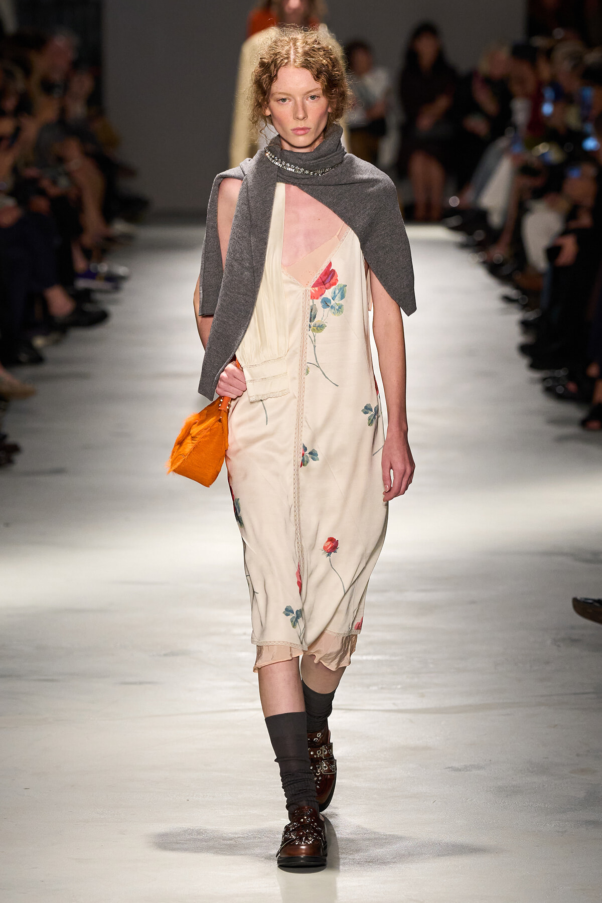 Female model walks the runway in a cream satin dress with floral embroidery, a gray cape, and an orange clutch.