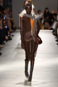 Model walks a fashion runway wearing a sheer brown dress with an orange panel, glittery shorts, and a gray scarf draped over one shoulder; accessorized with a brown handbag and chunky jewelry.