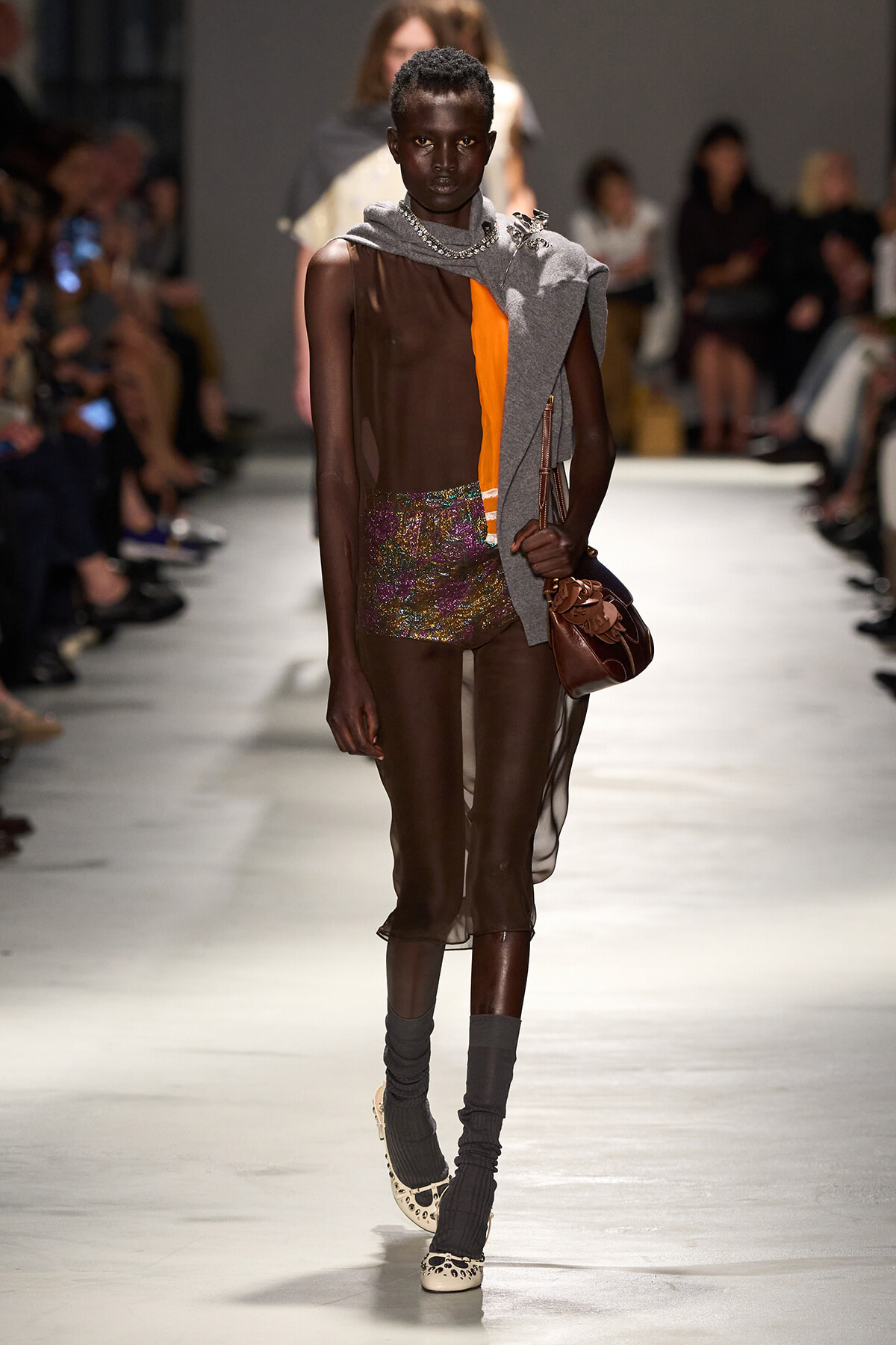 Model walks a fashion runway wearing a sheer brown dress with an orange panel, glittery shorts, and a gray scarf draped over one shoulder; accessorized with a brown handbag and chunky jewelry.