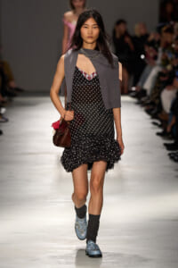 Model walking runway in a black polka-dot dress with ruffled hem, gray sleeveless cardigan, blue studded boots, and a brown bag.