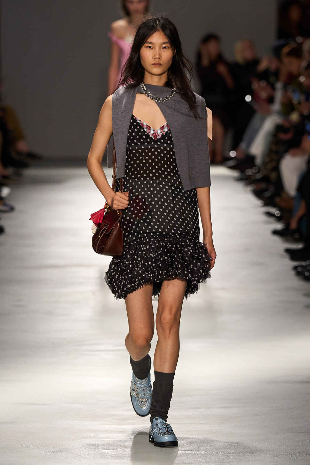 Model walking runway in a black polka-dot dress with ruffled hem, gray sleeveless cardigan, blue studded boots, and a brown bag.