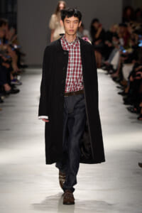 Male model on a runway wearing a long black coat over a red plaid shirt, dark jeans, and brown studded boots.
