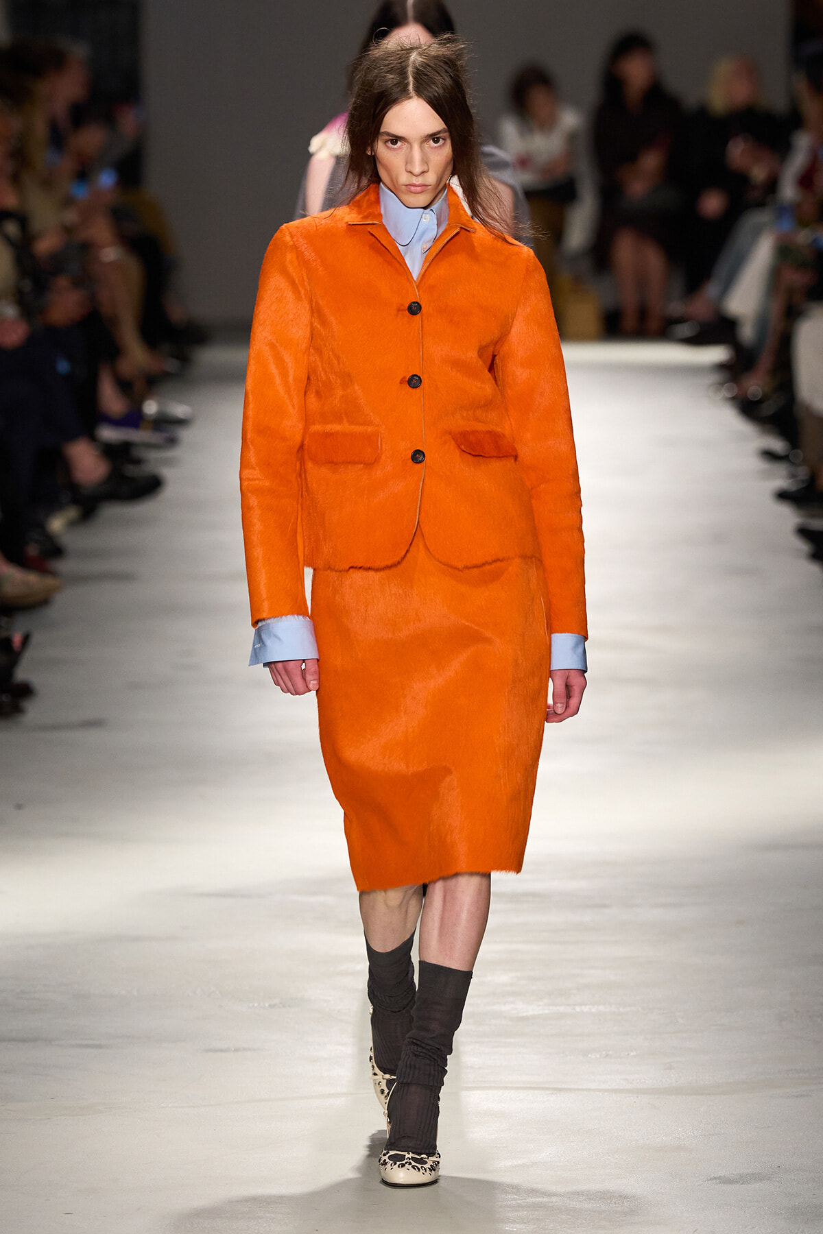Model walks the runway in a vivid orange suit with blue shirt cuffs visible at the wrists, pairing with leopard-printed shoes and dark socks.