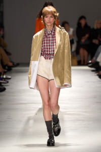 Model on a runway wearing a beige oversized coat over a red plaid shirt, cream high-waisted shorts, and black embellished loafers with dark socks.