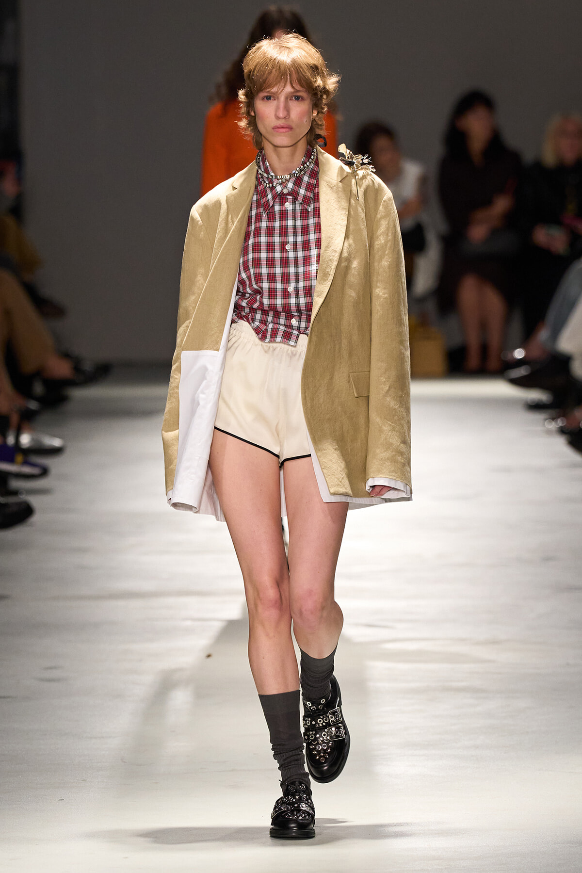 Model on a runway wearing a beige oversized coat over a red plaid shirt, cream high-waisted shorts, and black embellished loafers with dark socks.
