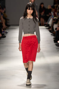 Model walks a fashion runway in a gray cardigan with a polka-dot collar, and a bright red pleated midi skirt, black socks, and patterned shoes.