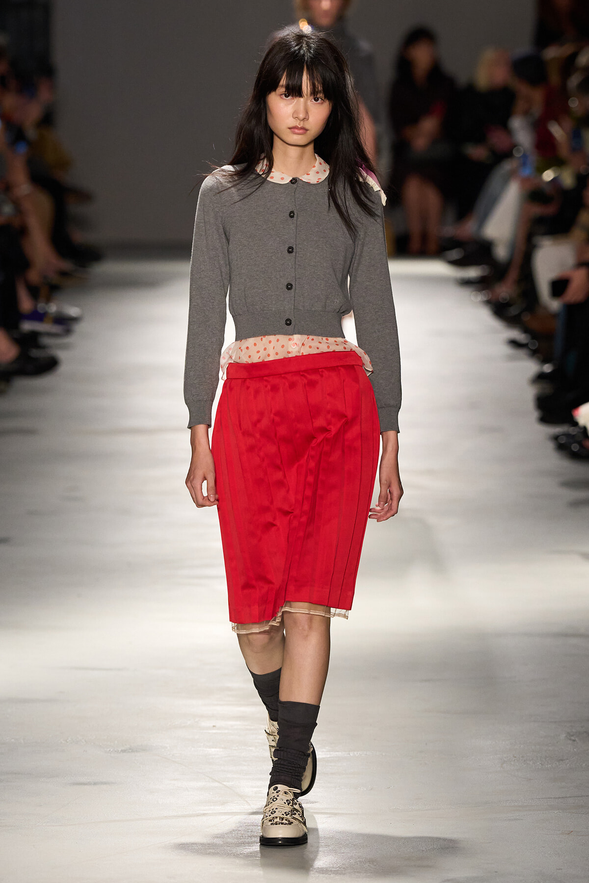Model walks a fashion runway in a gray cardigan with a polka-dot collar, and a bright red pleated midi skirt, black socks, and patterned shoes.