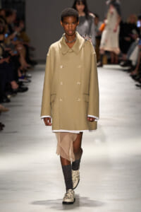 Model walking a runway in a tan oversized double-breasted coat over a white shirt and sheer beige skirt, with studded shoes.