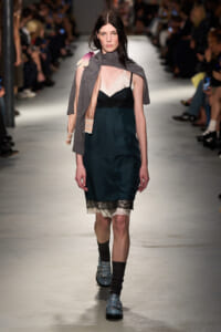 Model walks the runway in a teal satin slip dress with cream lace trim, layered under a pinkish slip, and a gray scarf draped over one shoulder.