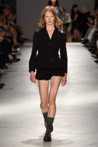 Model walks a runway in a black tailored blazer with a brooch and matching black shorts on a fashion show catwalk.