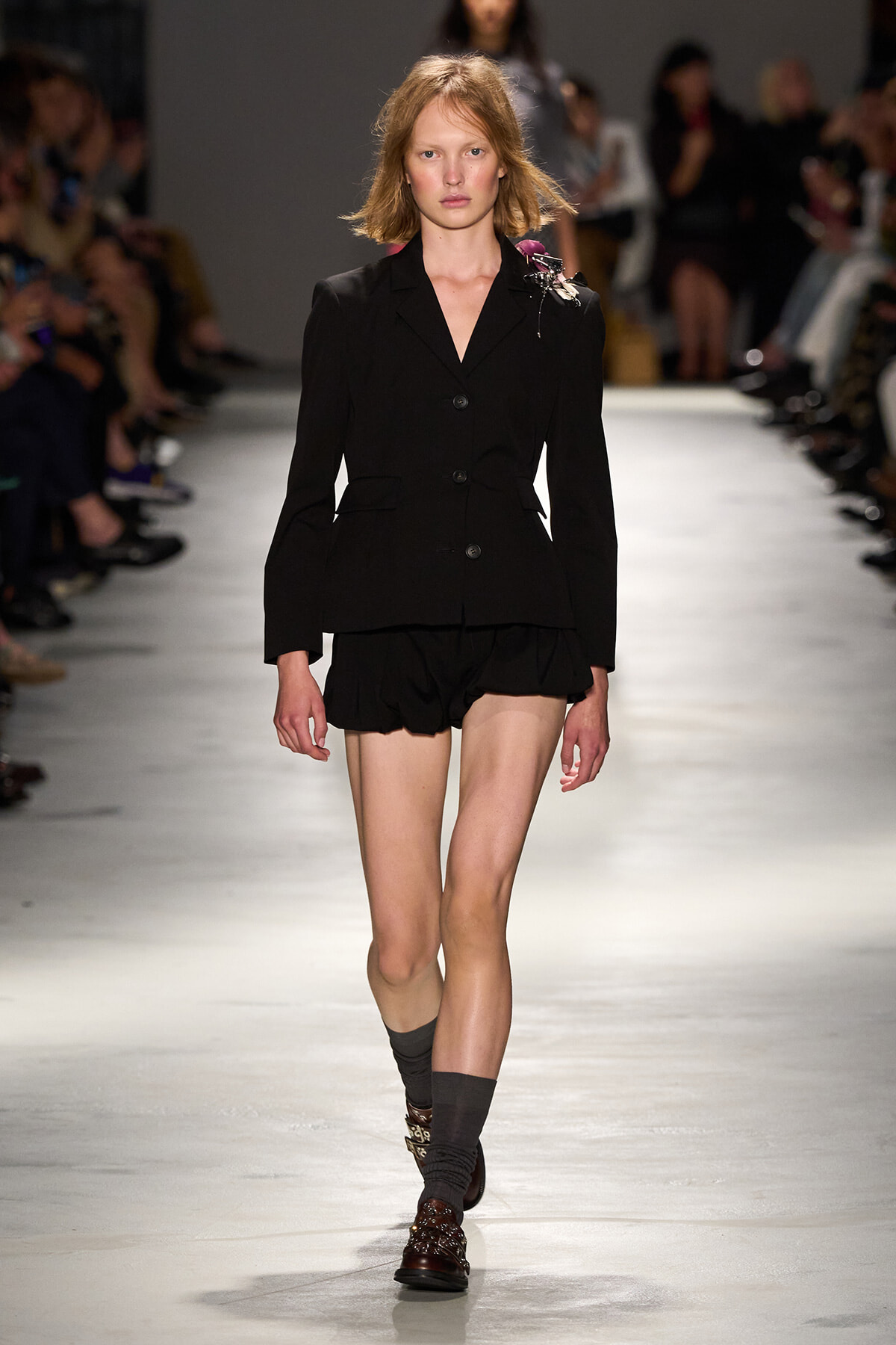 Model walks a runway in a black tailored blazer with a brooch and matching black shorts on a fashion show catwalk.