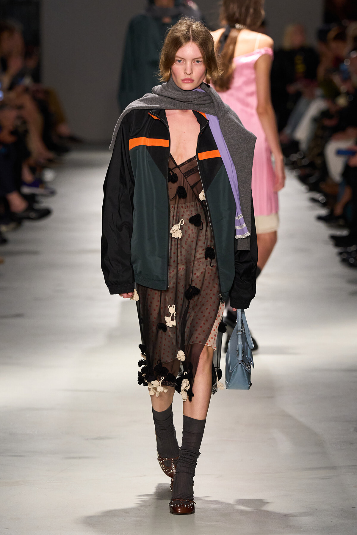 Model walking a fashion show wearing a color-blocked jacket over a sheer polka-dot dress with floral appliqués and a gray scarf, carrying a blue handbag.
