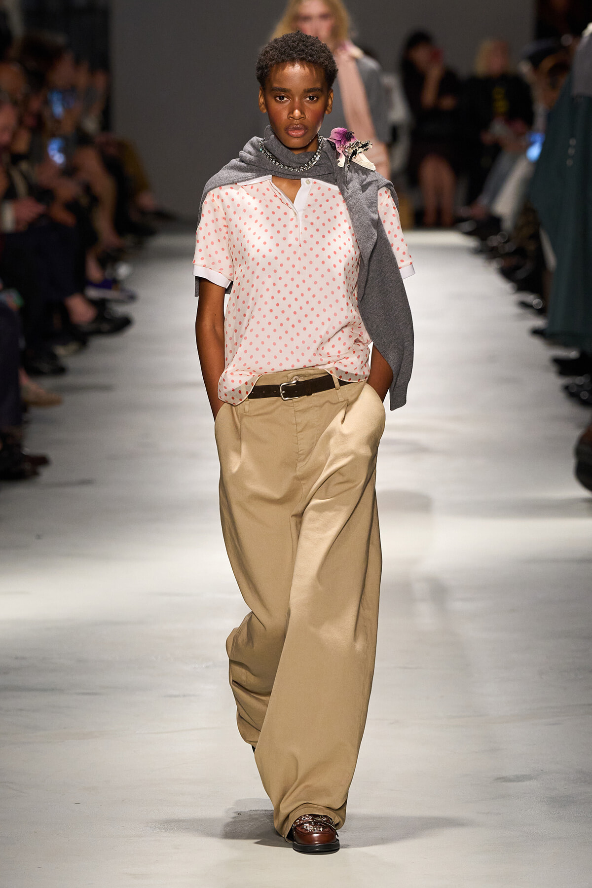 Model walking the runway in a pink polka-dot blouse with beige wide-leg trousers, brown belt, and a gray sweater draped over shoulders with a floral accessory at the neck.
